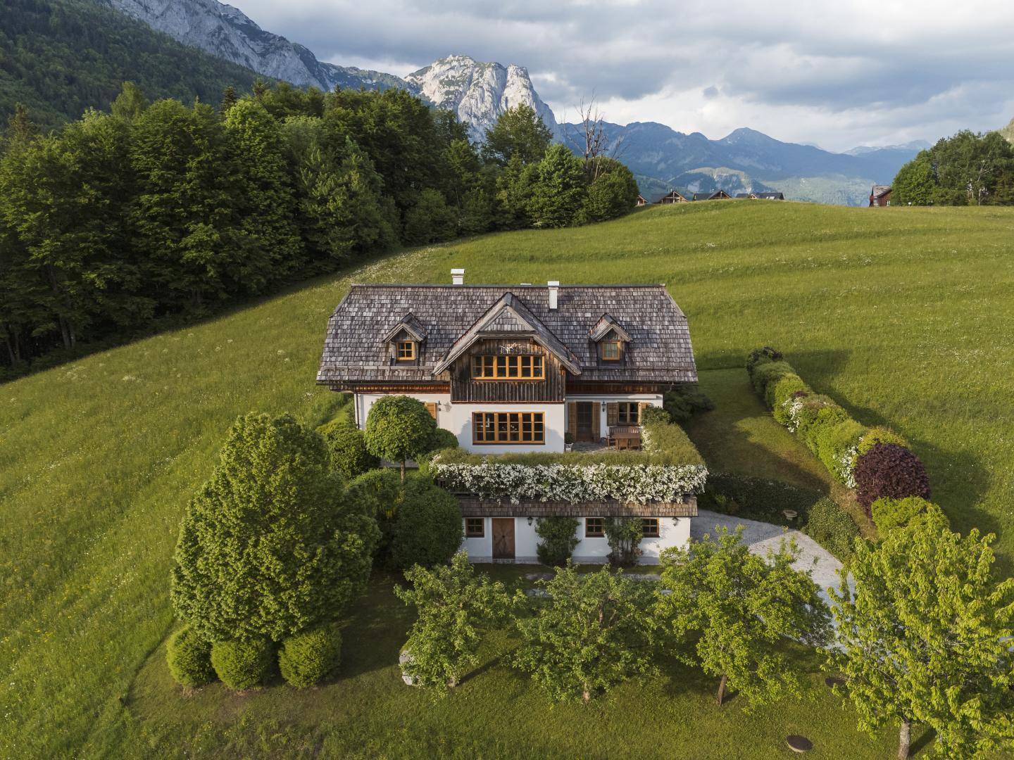 Panoramablick auf das charmante Landhaus mit üppiger grüner Umgebung und Berggipfeln im Hintergrund.