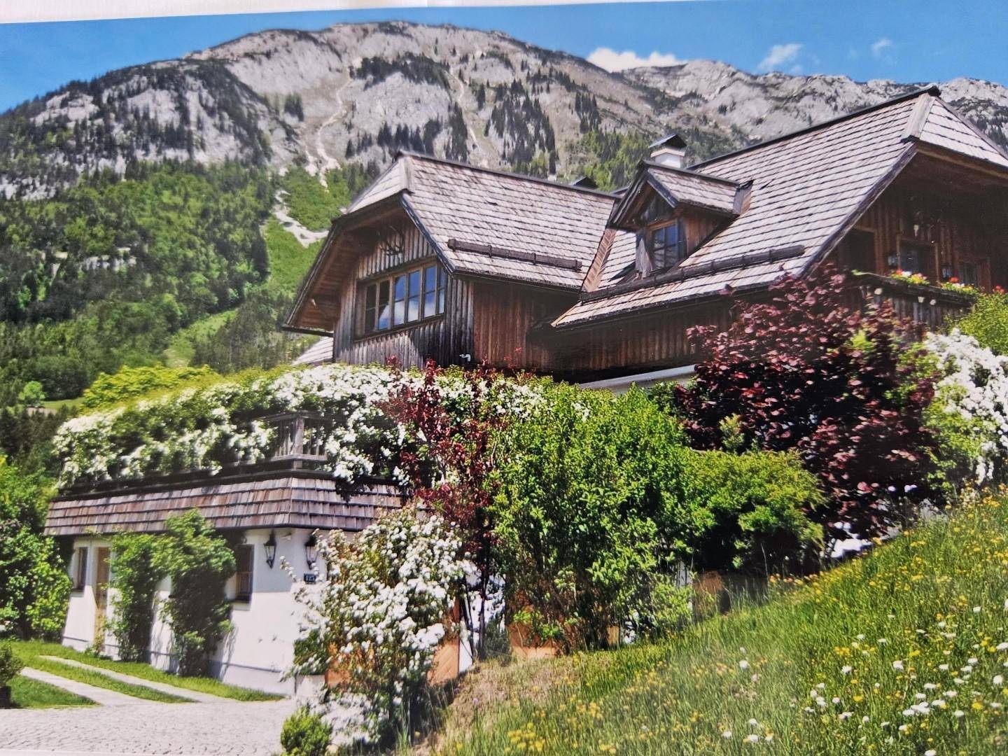 Detailansicht des Landhauses mit blühenden Sträuchern und traditioneller Holzfassade vor Bergkulisse.