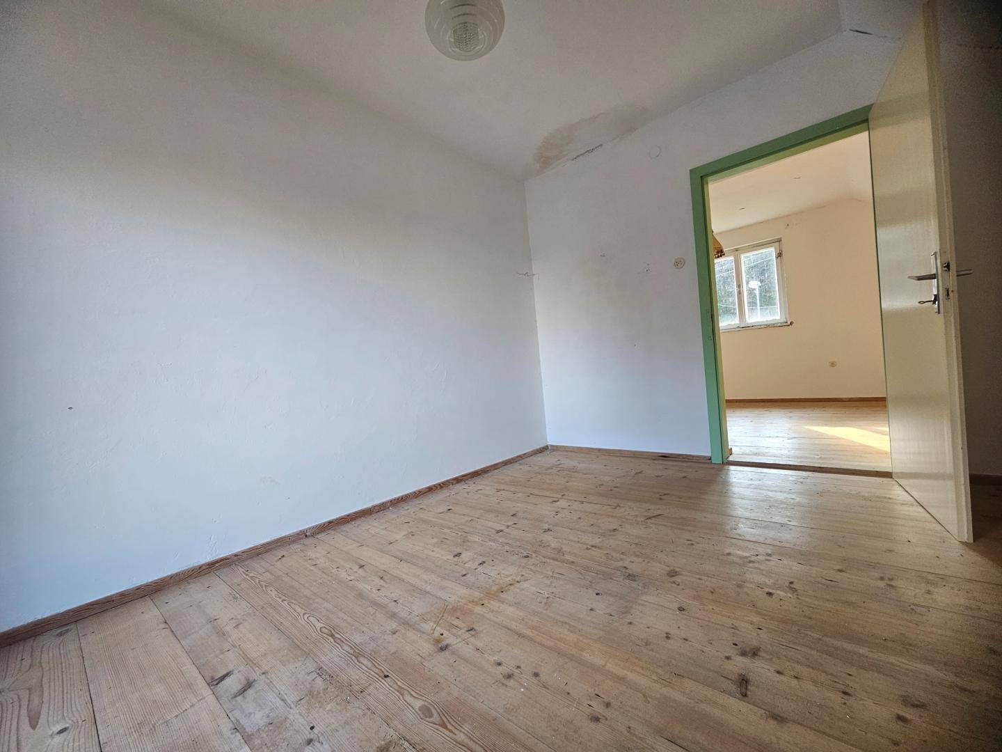 Heller, leerer Raum mit Holzdielenboden und weiß gestrichenen Wänden, mit Zugang zu einem weiteren Zimmer.