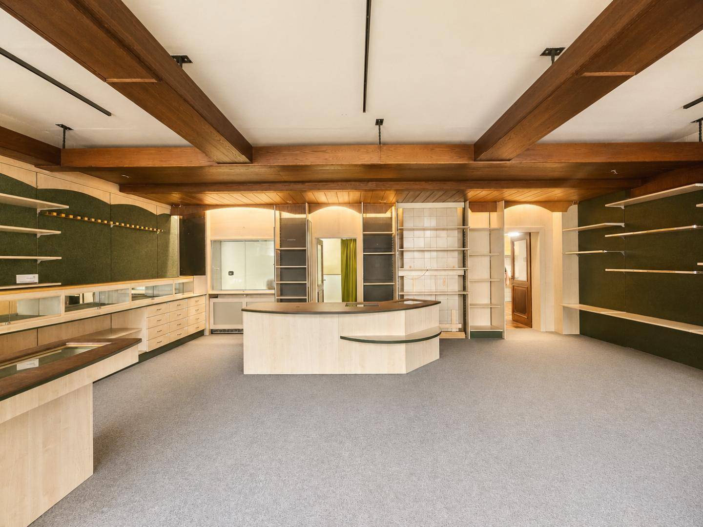 Großer Verkaufsraum mit hohen Decken, sichtbaren Holzbalken und Teppichboden, ausgestattet mit Regalen und Theke.