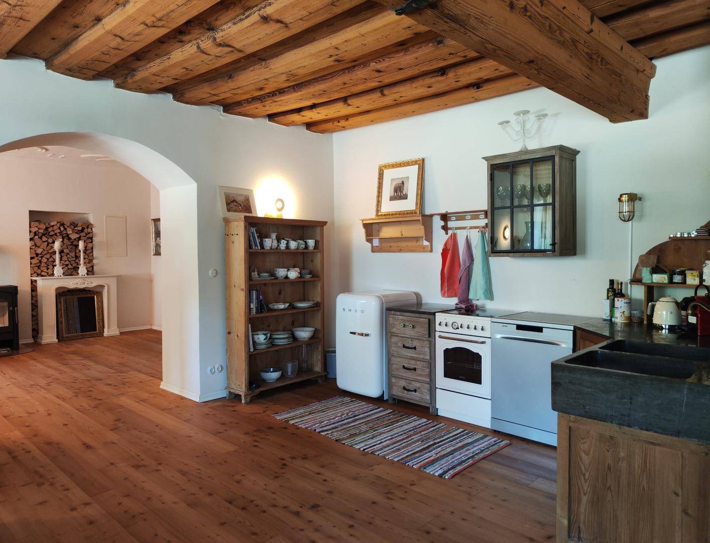 Rustikale Küche mit Holzbalkendecke, freistehendem Kühlschrank und Blick in den Wohnbereich.