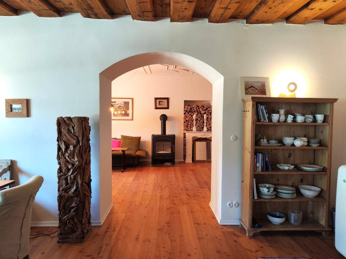 Blick durch einen Rundbogen in ein gemütliches Wohnzimmer mit Kaminofen und Holzregal.