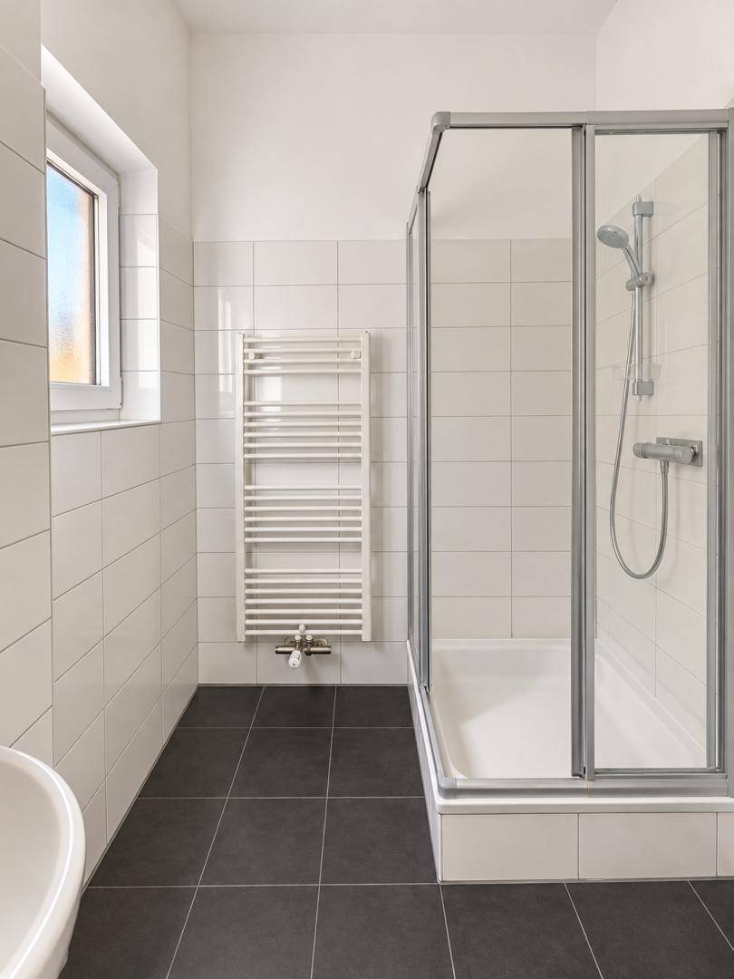 Modernes Badezimmer mit Duschkabine, weißen Wandfliesen, dunklem Boden und Fenster.