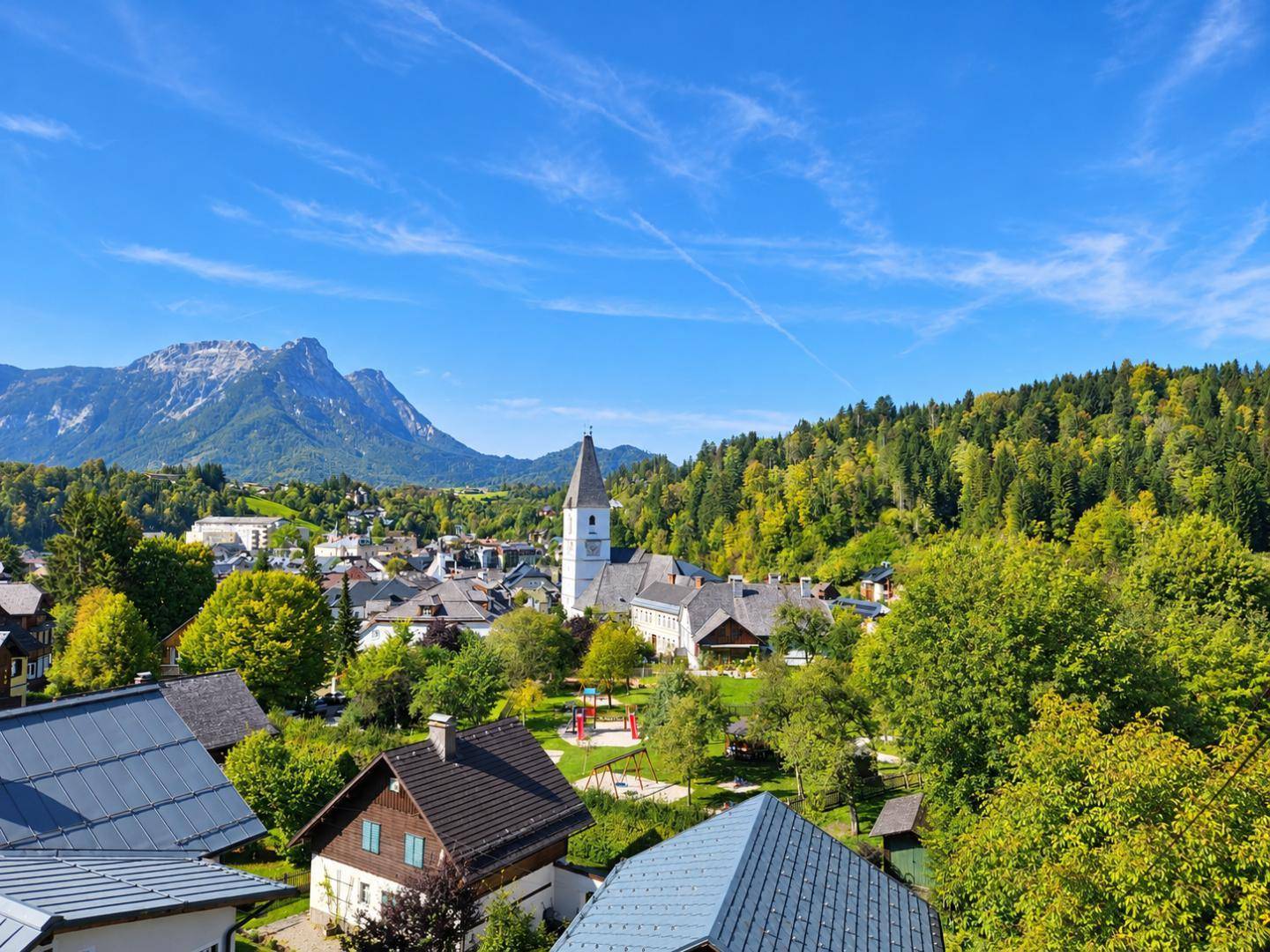 Malerischer Blick über eine Stadt mit Kirche, umgeben von grünen Hügeln und Bergen.