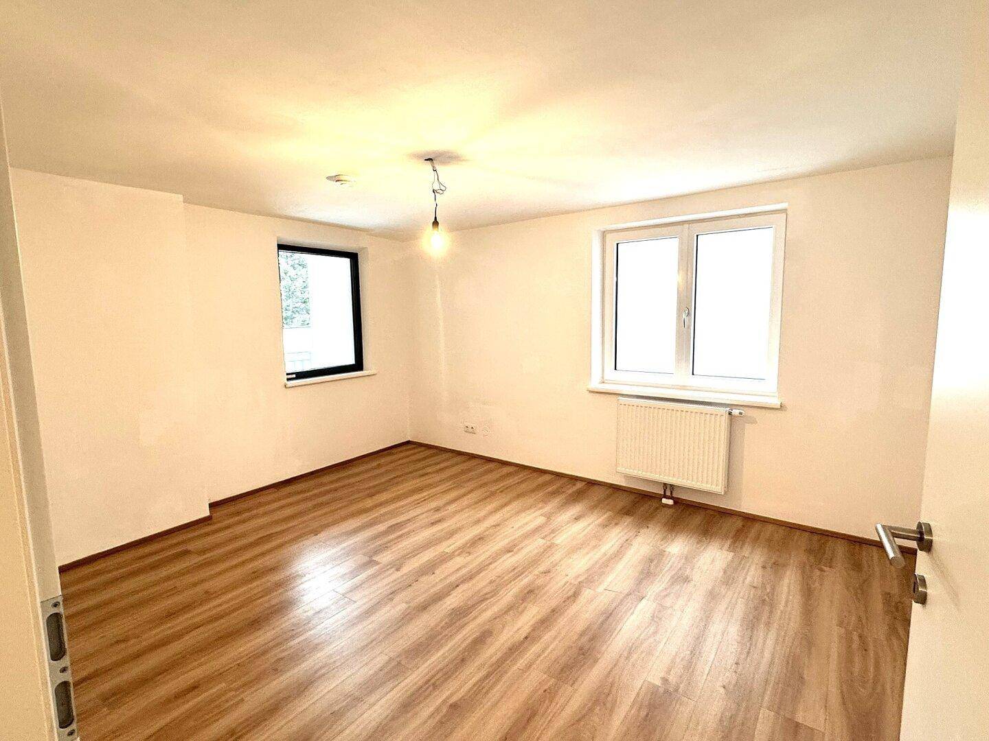 Geräumiger, heller Raum mit Laminatboden und zwei Fenstern, eines davon mit dunklem Rahmen.
