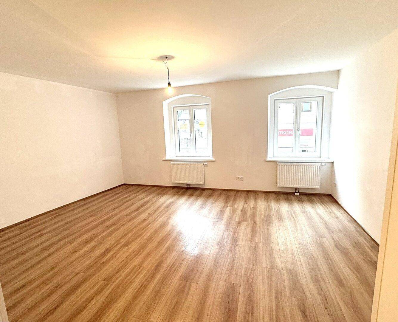 Heller, leerer Raum mit zwei Fenstern, Laminatboden und weiß gestrichenen Wänden.