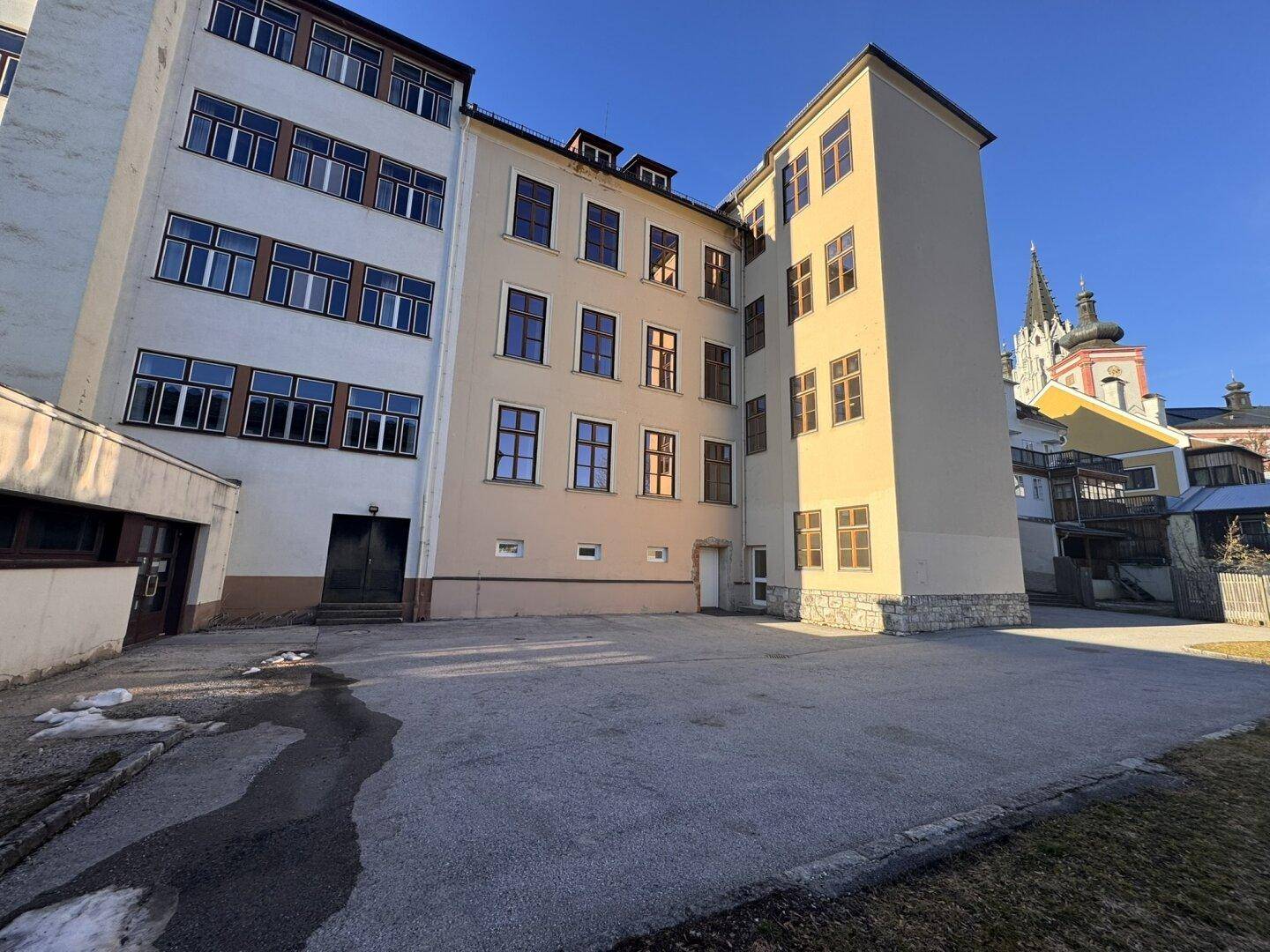 Seitenansicht eines großen Gebäudes mit asphaltiertem Vorplatz und einem Kirchturm im Hintergrund.