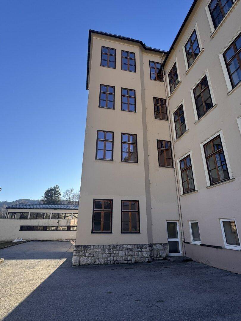 Seitenfassade eines Gebäudes mit vielen Fenstern und einem gepflasterten Bereich davor.