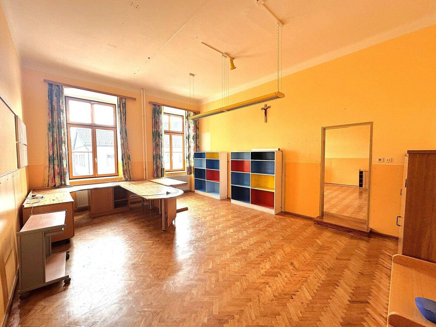 Großer Raum mit Fischgrätparkett, hohen Decken und bunten Regalen, ideal als Büro oder Klassenzimmer.