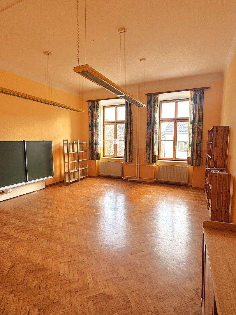 Heller Raum mit Fischgrätparkett, großen Fenstern und Regalen, geeignet als Büro oder Lernbereich.