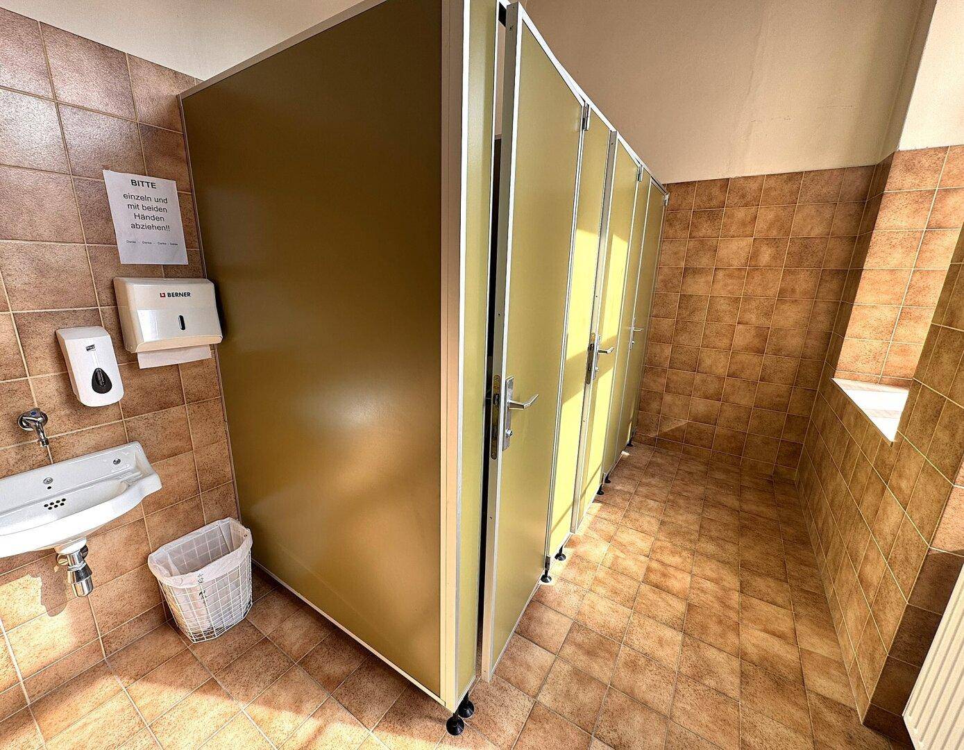 Sanitärbereich mit mehreren Toilettenkabinen, Fliesenboden und einem Waschbecken.