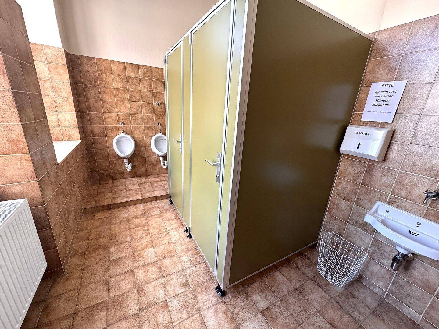 Toilettenraum mit mehreren Kabinen, Urinalen und einem Waschbecken, gefliest in Brauntönen.