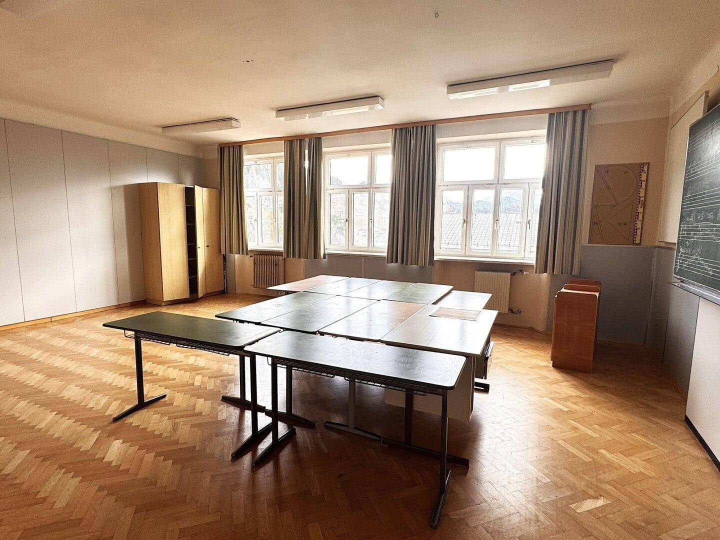 Heller Raum mit Holzboden, mehreren Tischen und großen Fenstern mit Vorhängen.
