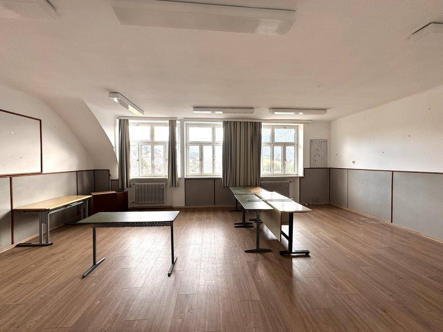 Heller Raum mit Laminatboden, Tischen und großen Fenstern mit Blick ins Grüne.