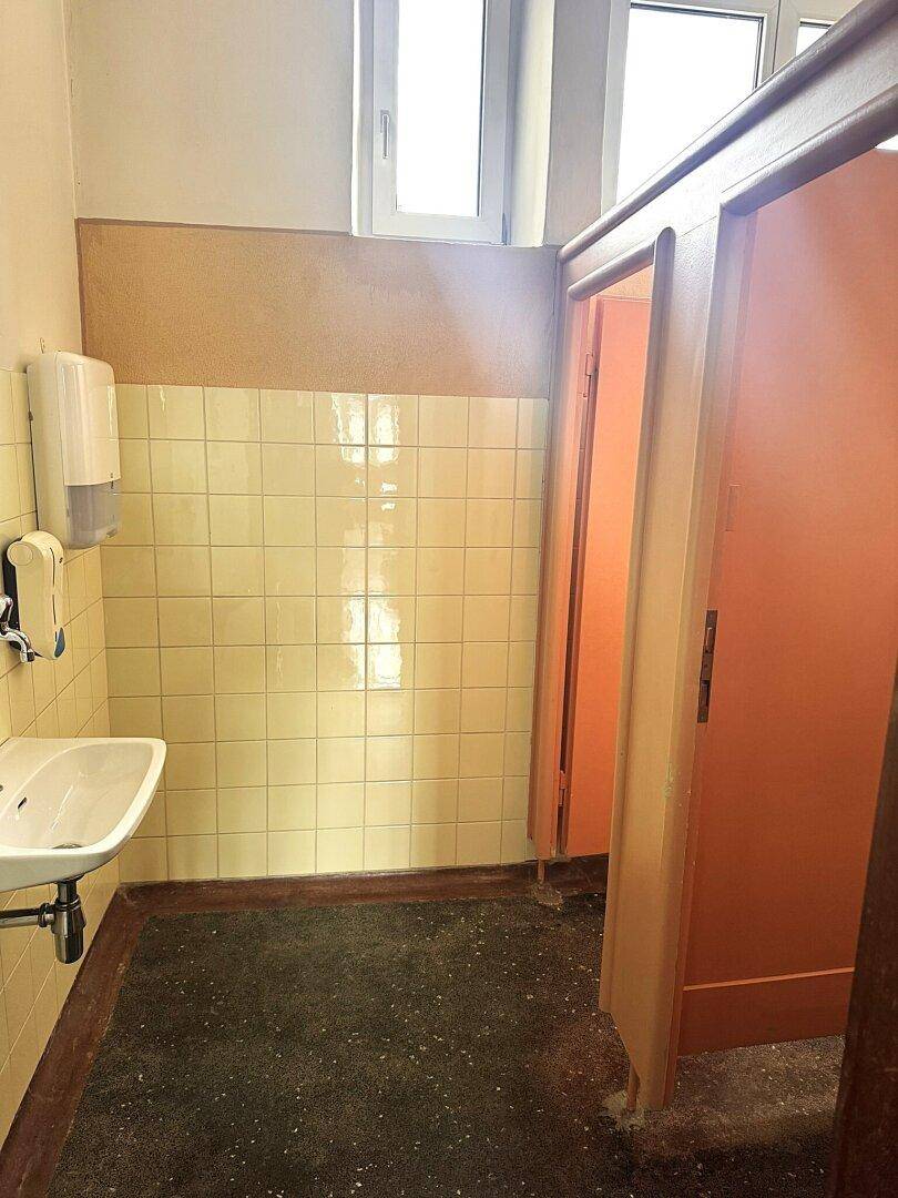 Öffentliche Toilette mit gefliesten Wänden, Waschbecken und orangefarbenen Kabinentüren.