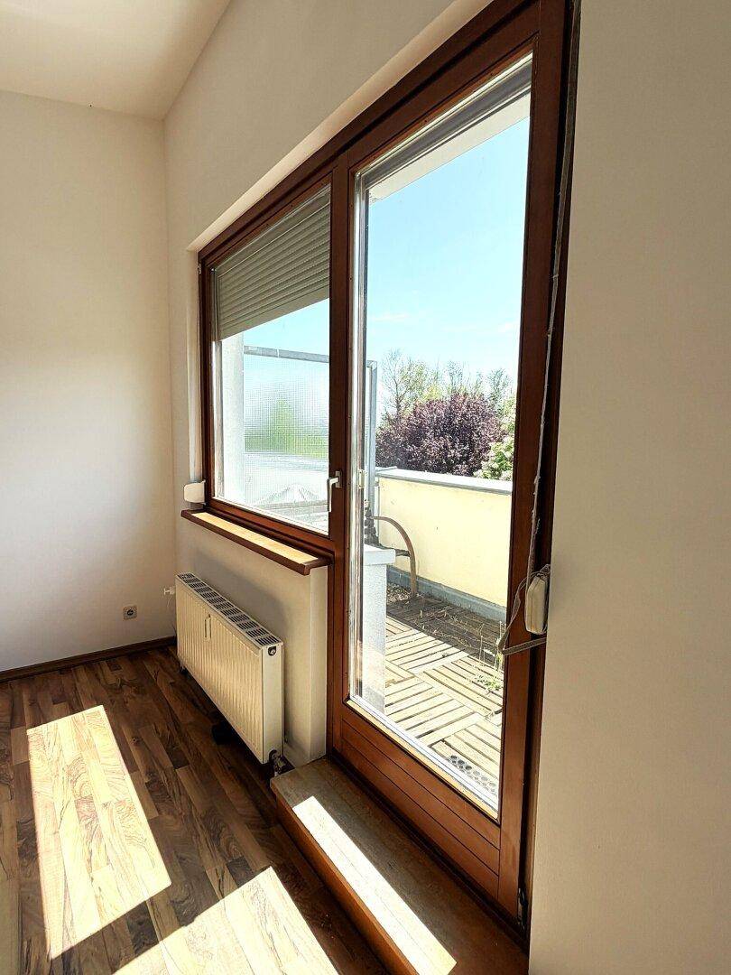 Heller Raum mit Holzboden und großer Glastür, die zu einem sonnigen Außenbereich führt.