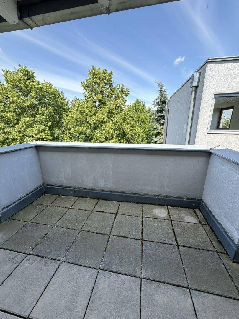 Leerer Balkon mit grauen Fliesen und Wänden, umgeben von grünen Bäumen im Hintergrund.