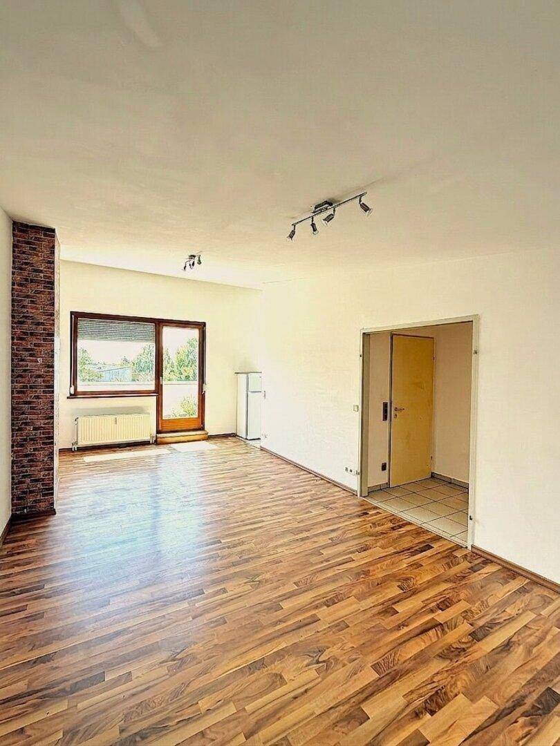 Geräumiges, leeres Wohnzimmer mit Holzboden, Ziegelwand und Zugang zum Außenbereich.