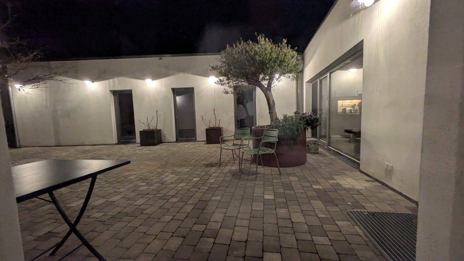 Beleuchteter gepflasterter Innenhof bei Nacht mit Sitzgelegenheiten und einem zentralen Olivenbaum.