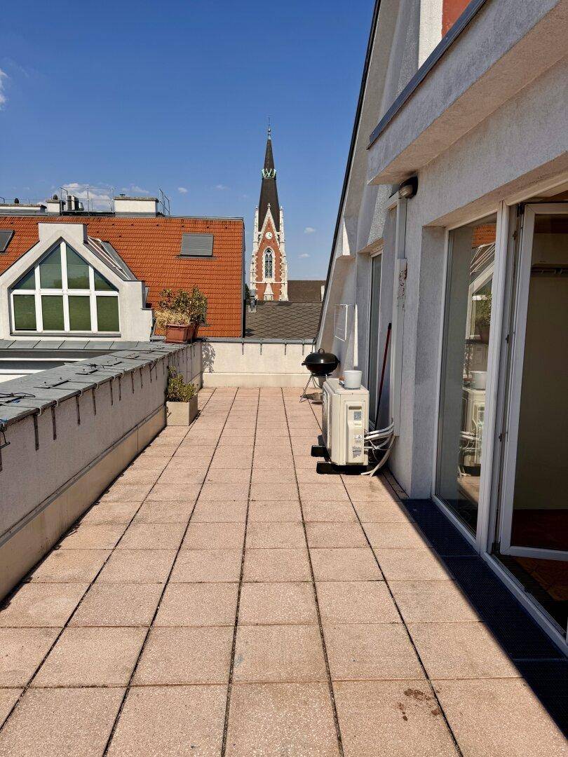 Sonnige Dachterrasse mit Zugang zum Innenbereich, Grill und Blick auf eine Kirchturmspitze.
