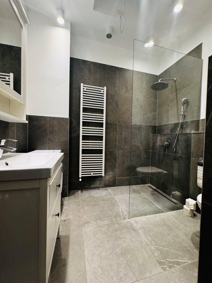 Elegantes Badezimmer mit Einzelwaschbecken, begehbarer Dusche und dunklen Fliesen.