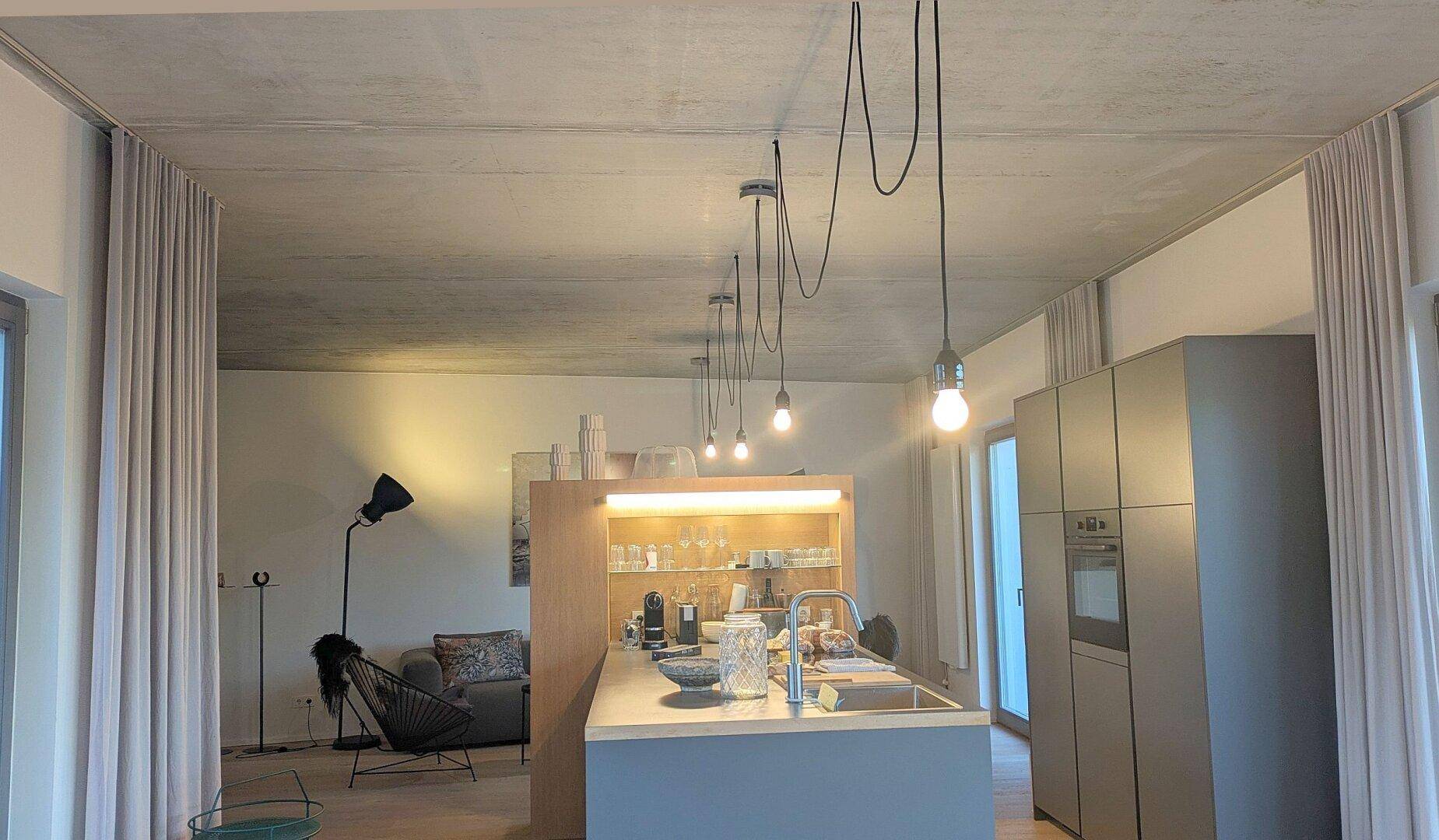 Offener Wohnbereich mit Kücheninsel, minimalistischen Hängelampen und Blick auf den Wohnbereich.