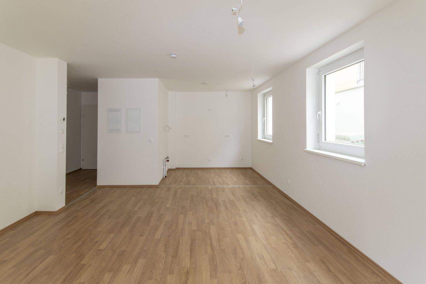 Geräumiger Wohnbereich mit hellem Laminatboden und mehreren Fenstern, die viel Licht hereinlassen.
