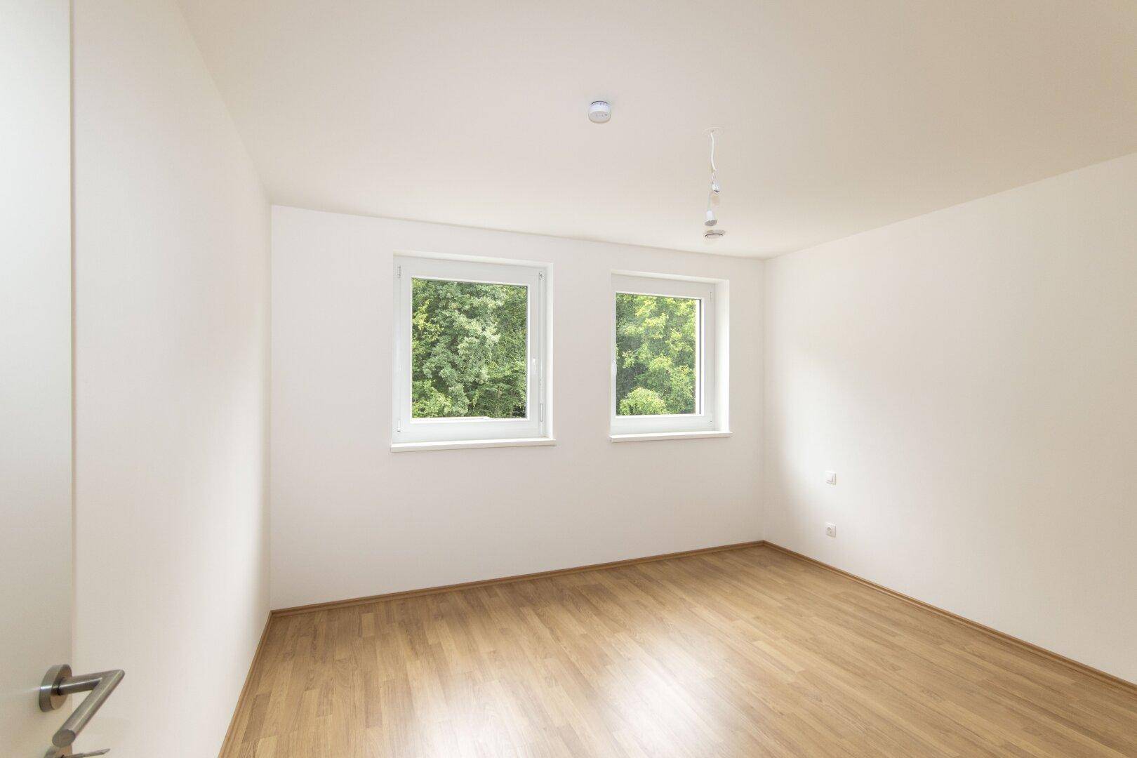 Helles Schlafzimmer mit Laminatboden und zwei Fenstern, die einen Blick ins Grüne bieten.