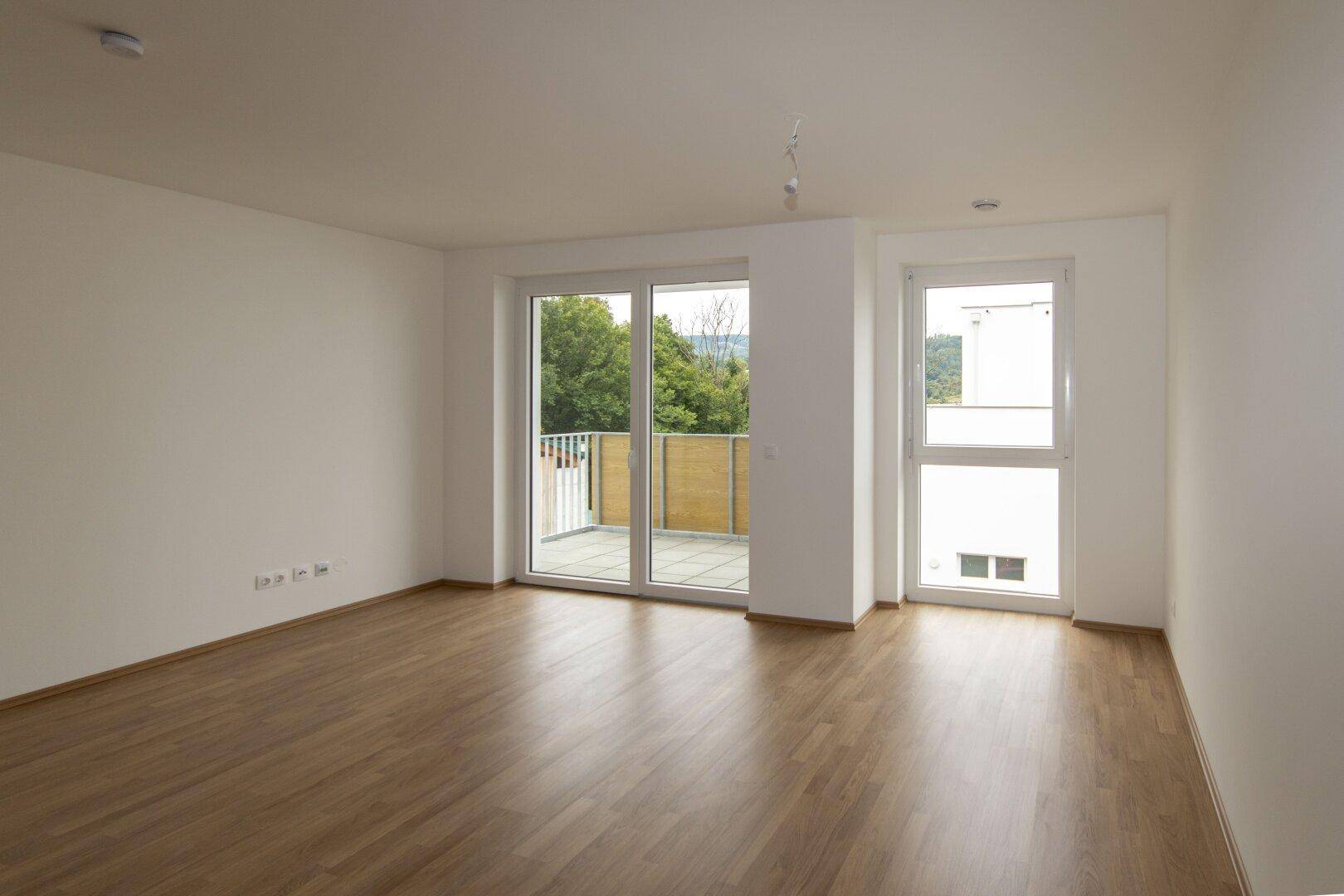 Geräumiges, leeres Wohnzimmer mit Laminatboden und Zugang zu einem Balkon.