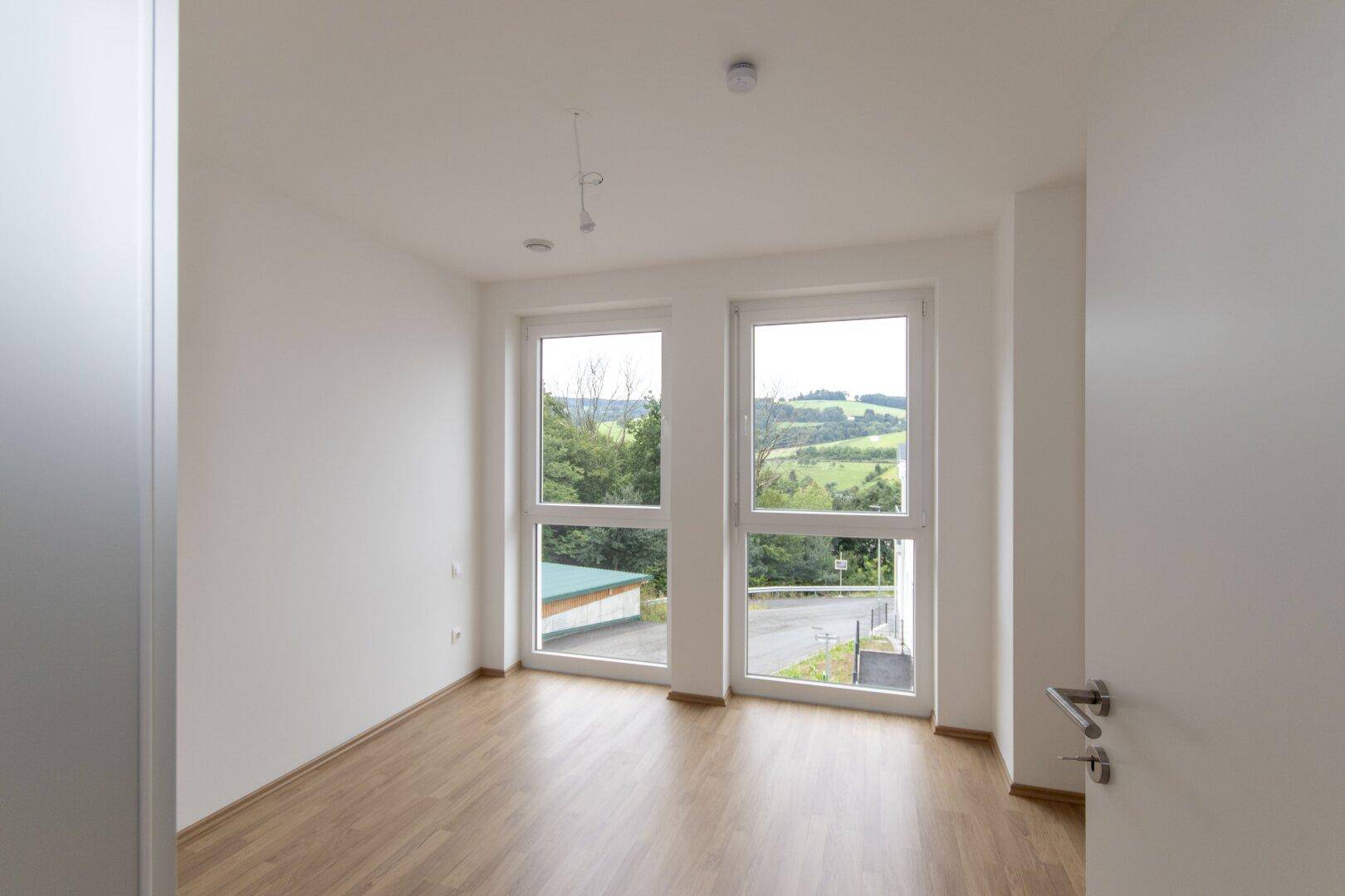 Helles Schlafzimmer mit Laminatboden und großen Fenstern mit Blick ins Grüne.