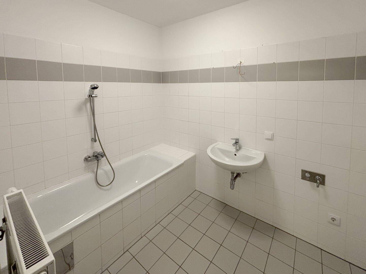 Badezimmer mit Badewanne, Waschbecken und weißen Fliesen, akzentuiert durch eine graue Bordüre.