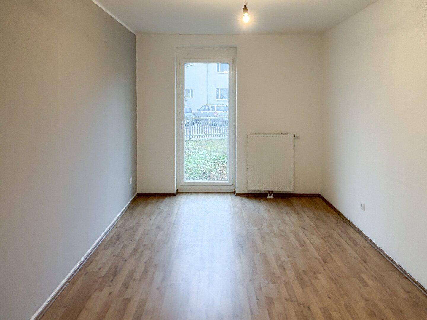 Leerer Raum mit Laminatboden, einer grauen Akzentwand und einem Fenster mit Blick ins Grüne.