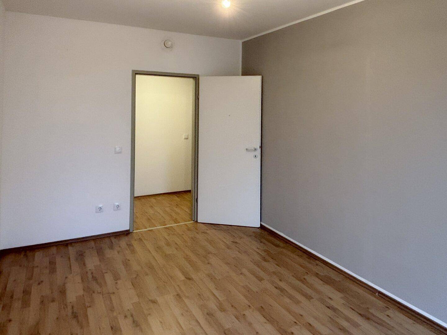 Unmöblierter Raum mit Laminatboden, einer grauen Wand und einer weißen Tür zu einem weiteren Zimmer.