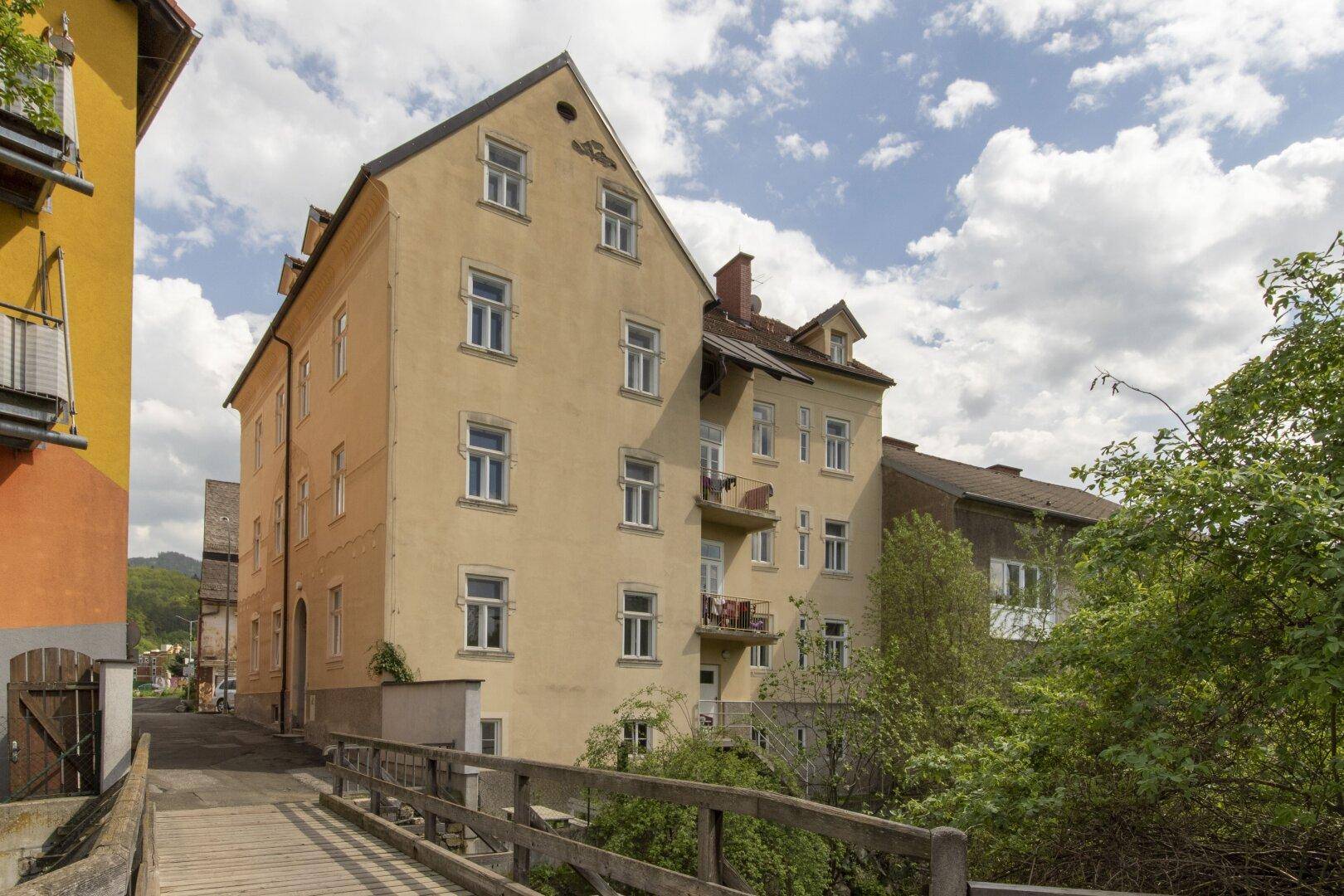 Historisches Mehrfamilienhaus mit heller Fassade, kleinen Balkonen und einer charmanten Holzbrücke im Vordergrund.