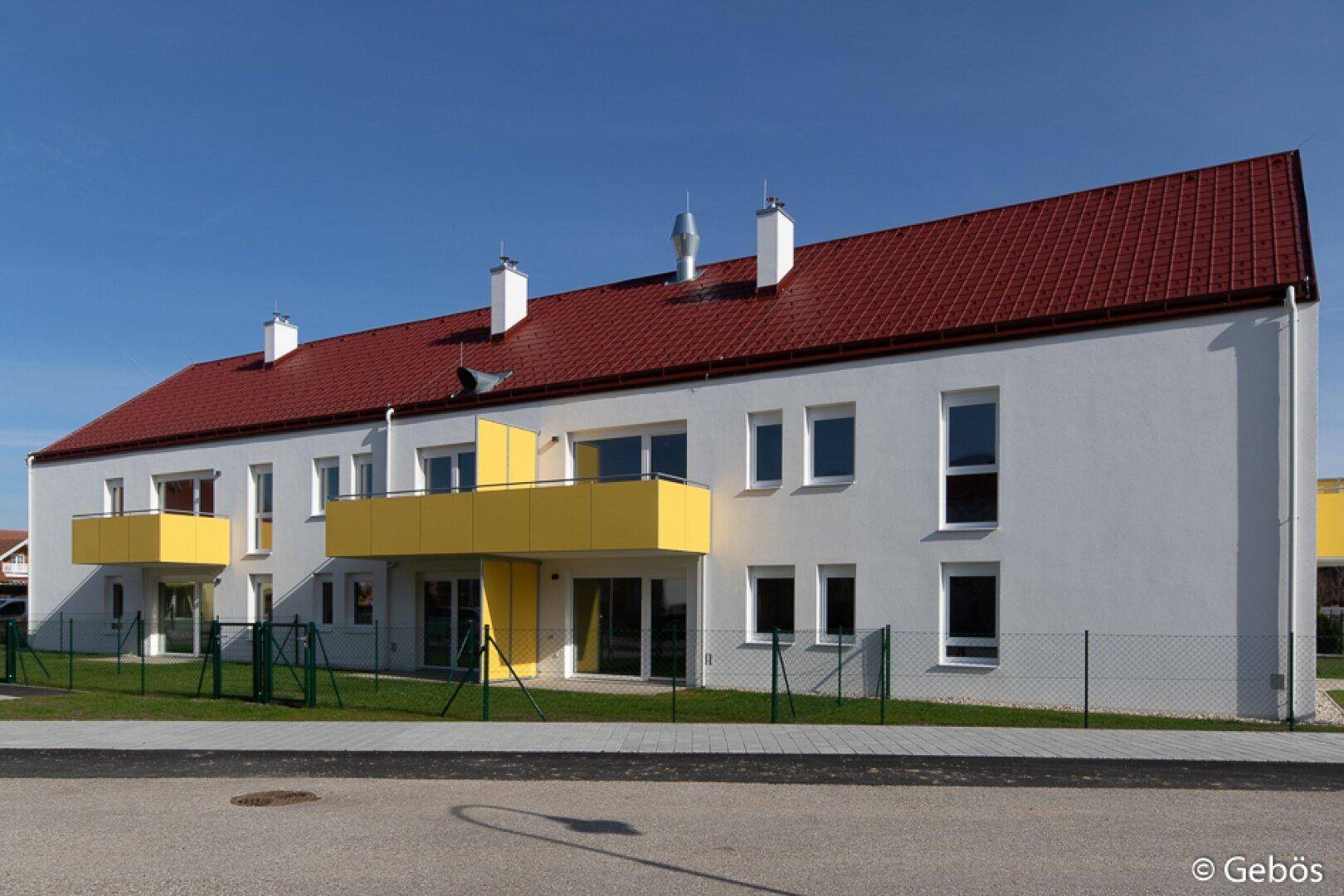 Moderne Wohnanlage mit weißer Fassade, rotem Satteldach und gelben Balkonen.