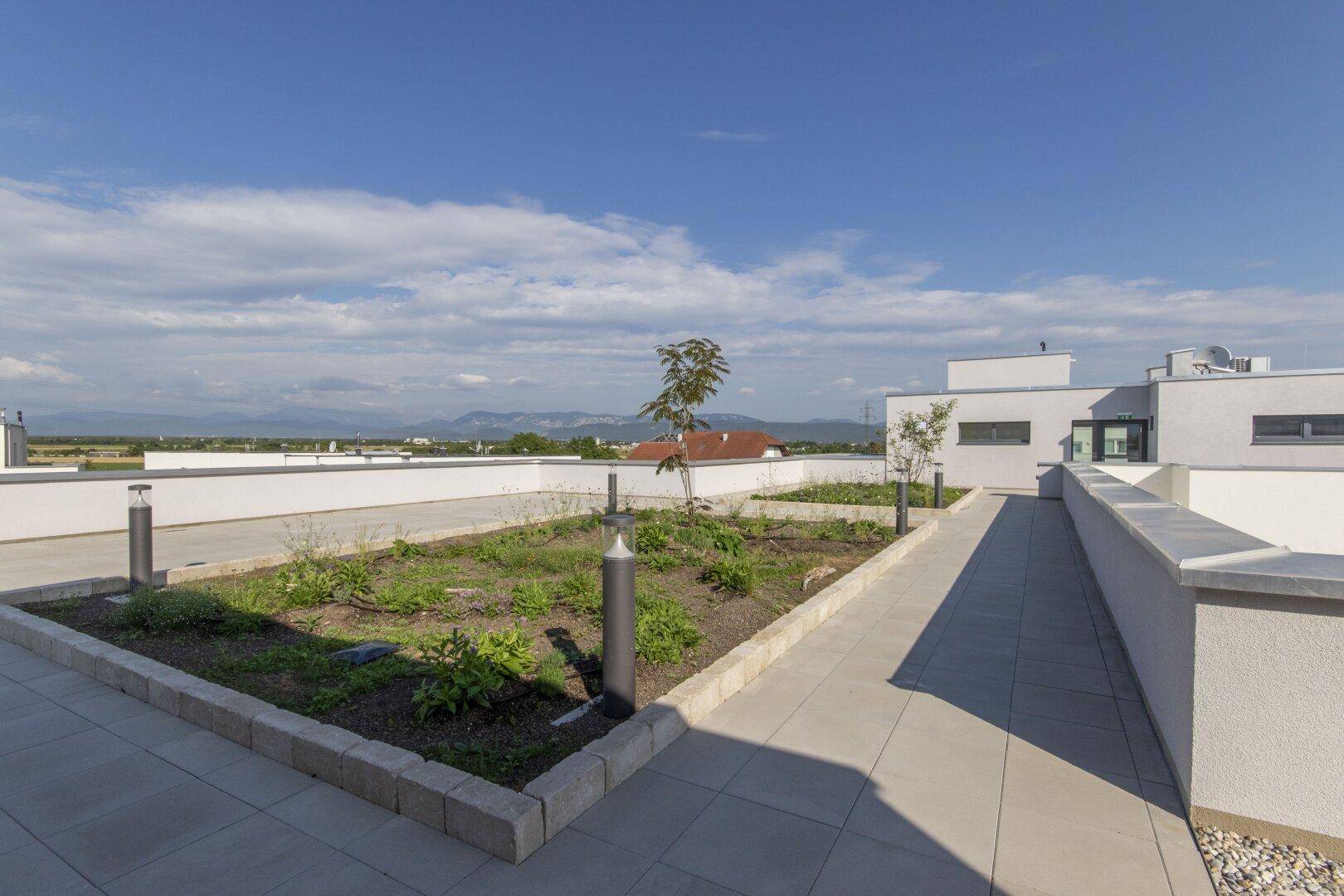 Moderne Dachterrasse mit begrünten Beeten, gepflasterten Wegen und weitem Blick auf die Landschaft und Berge.
