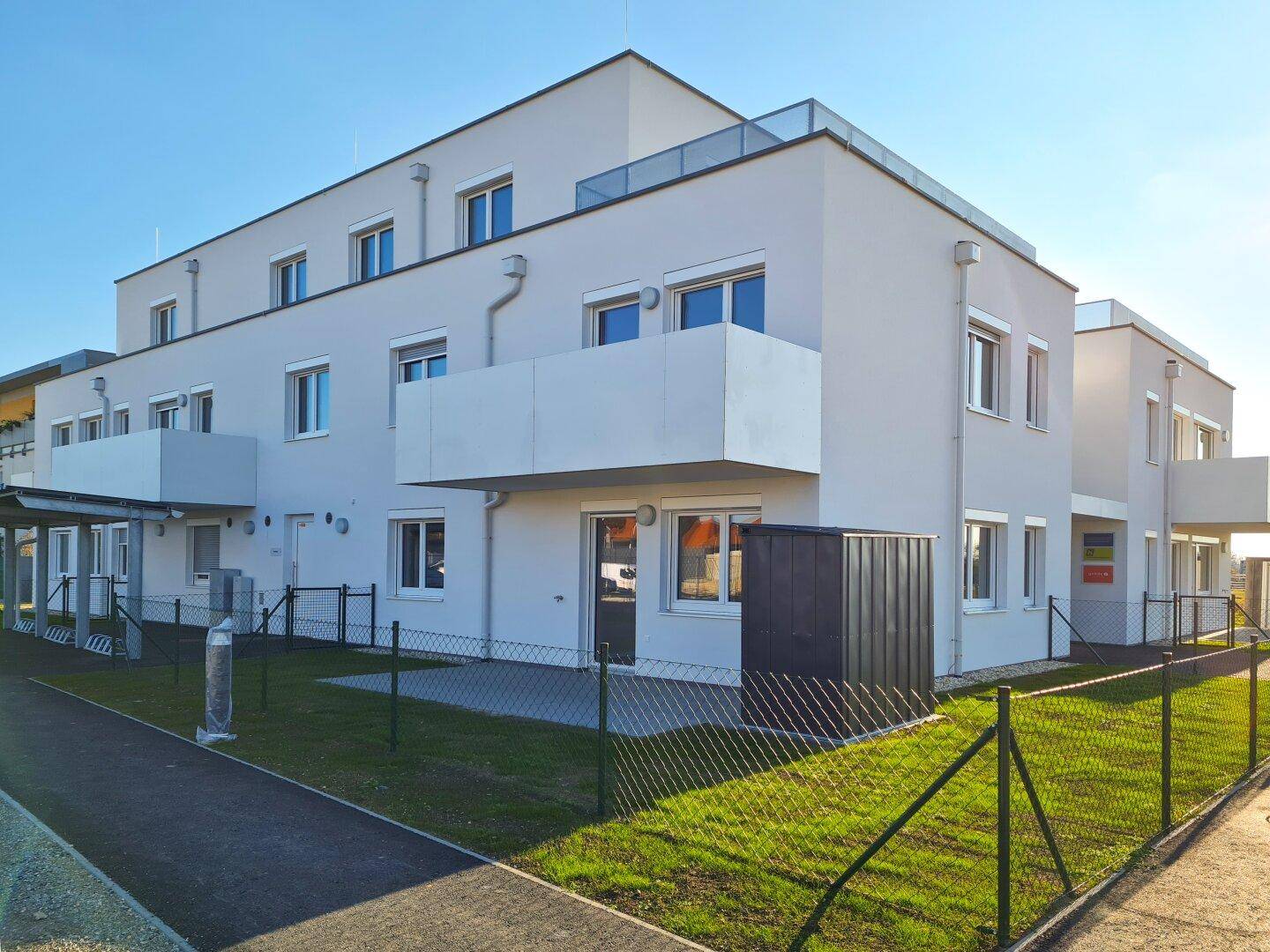 Modernes Mehrfamilienhaus mit heller Fassade, Balkonen, Dachterrasse und kleinen Gartenbereichen im Erdgeschoss.