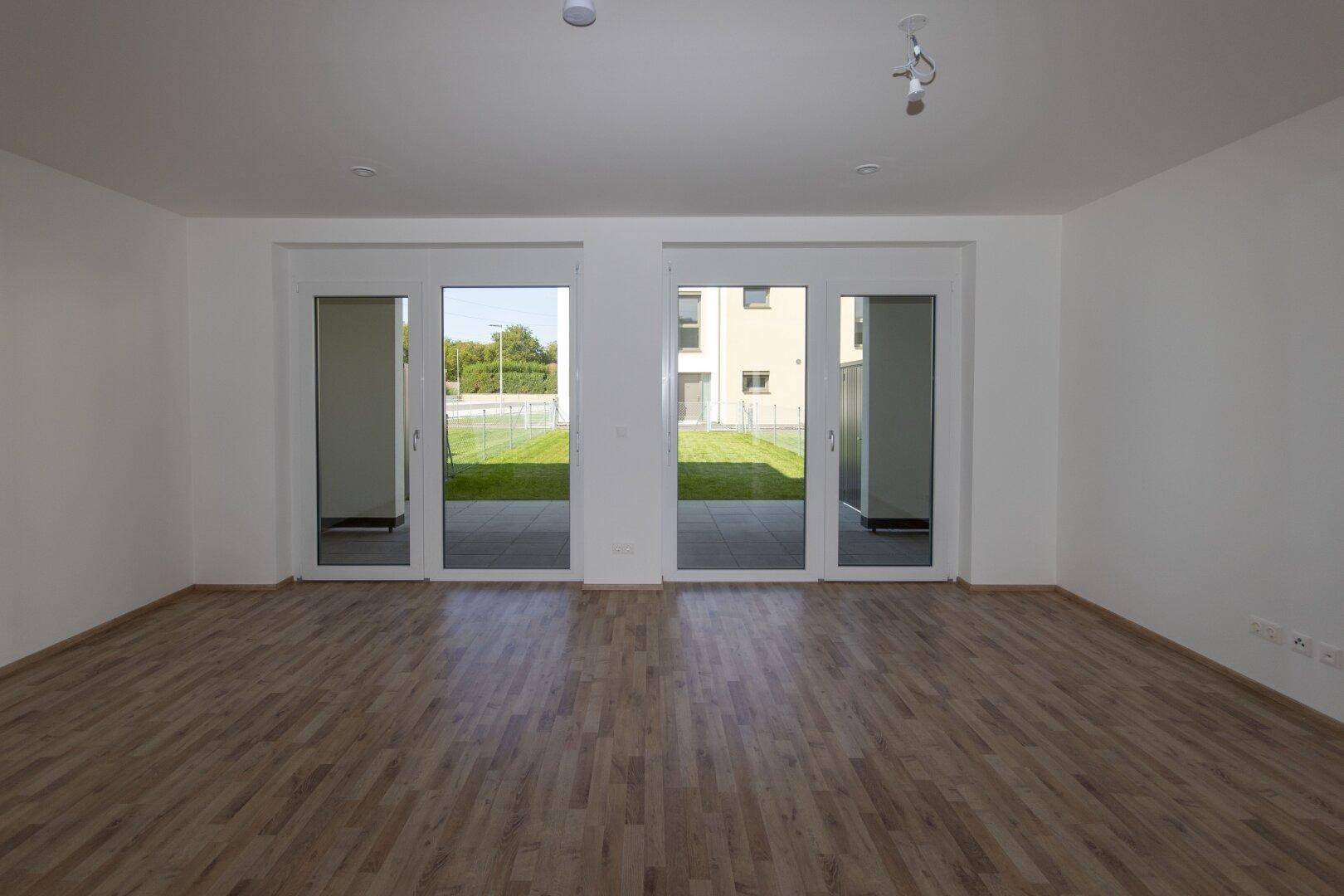 Heller, leerer Wohnraum mit Holzboden und großen Fenstertüren zum Garten.