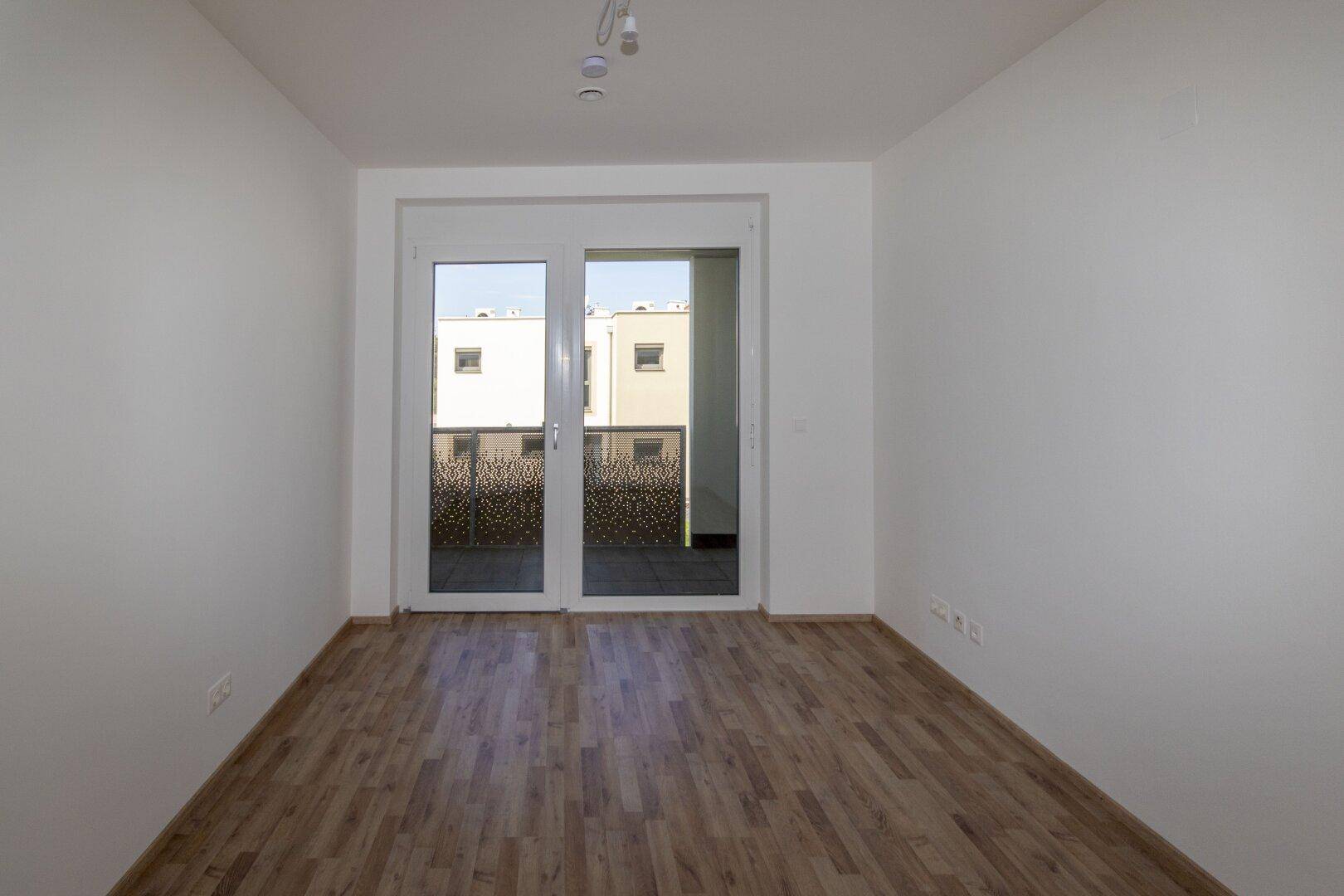 Geräumiges Zimmer mit Holzboden und großen Fenstertüren, die auf einen Balkon führen.