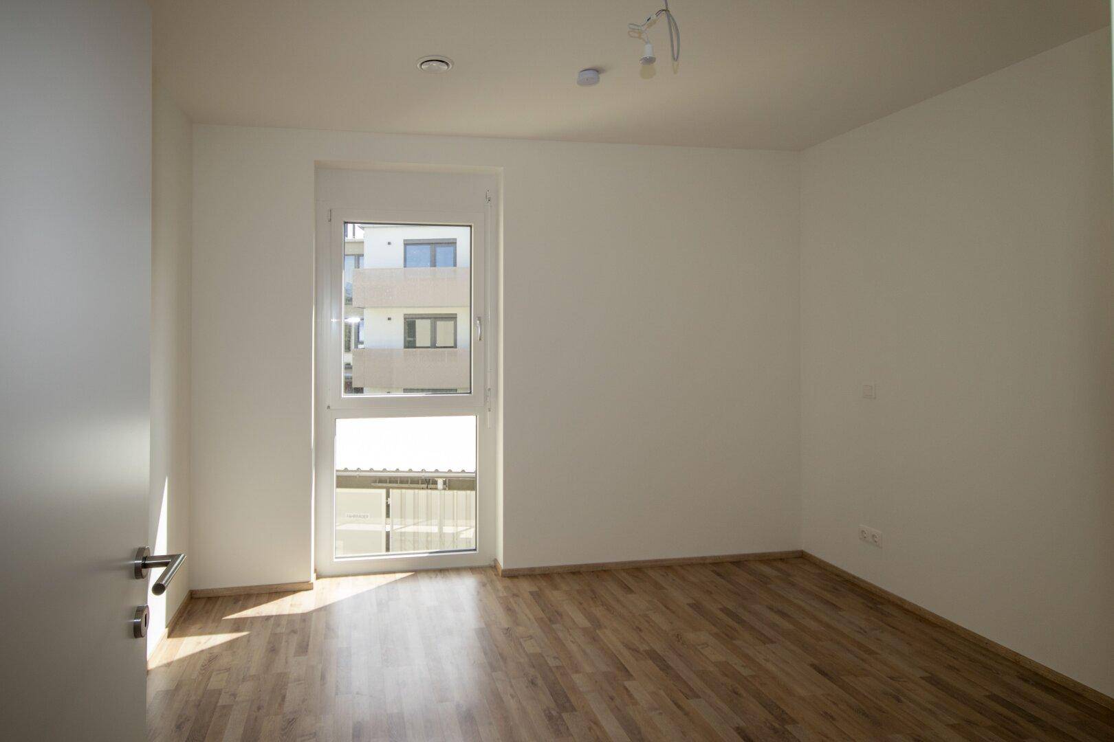 Sonniger Raum mit Holzboden und großem Fenster, das viel Tageslicht hereinlässt.