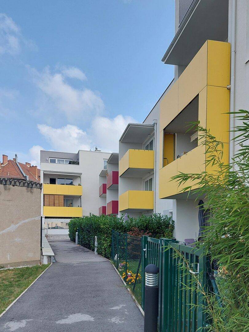 Ansicht einer modernen Wohnanlage mit farbigen Balkonen und einem begrünten Weg.