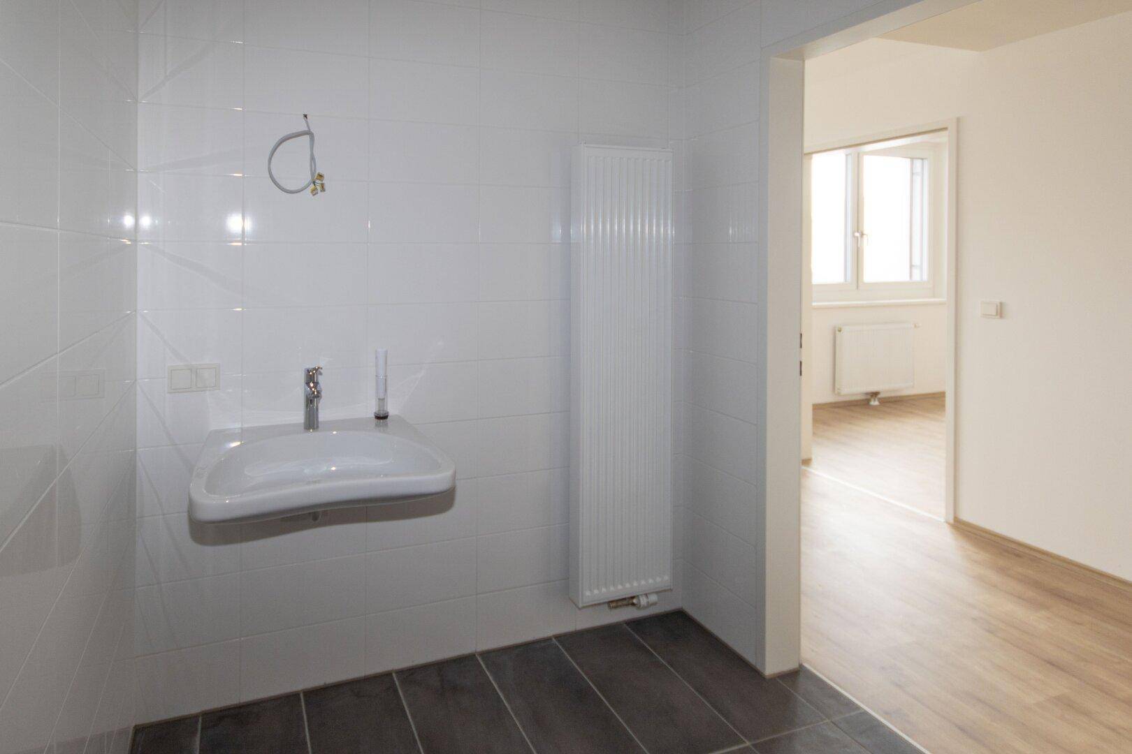 Helles Badezimmer mit Waschbecken, Handtuchheizkörper und Blick in einen angrenzenden Raum.