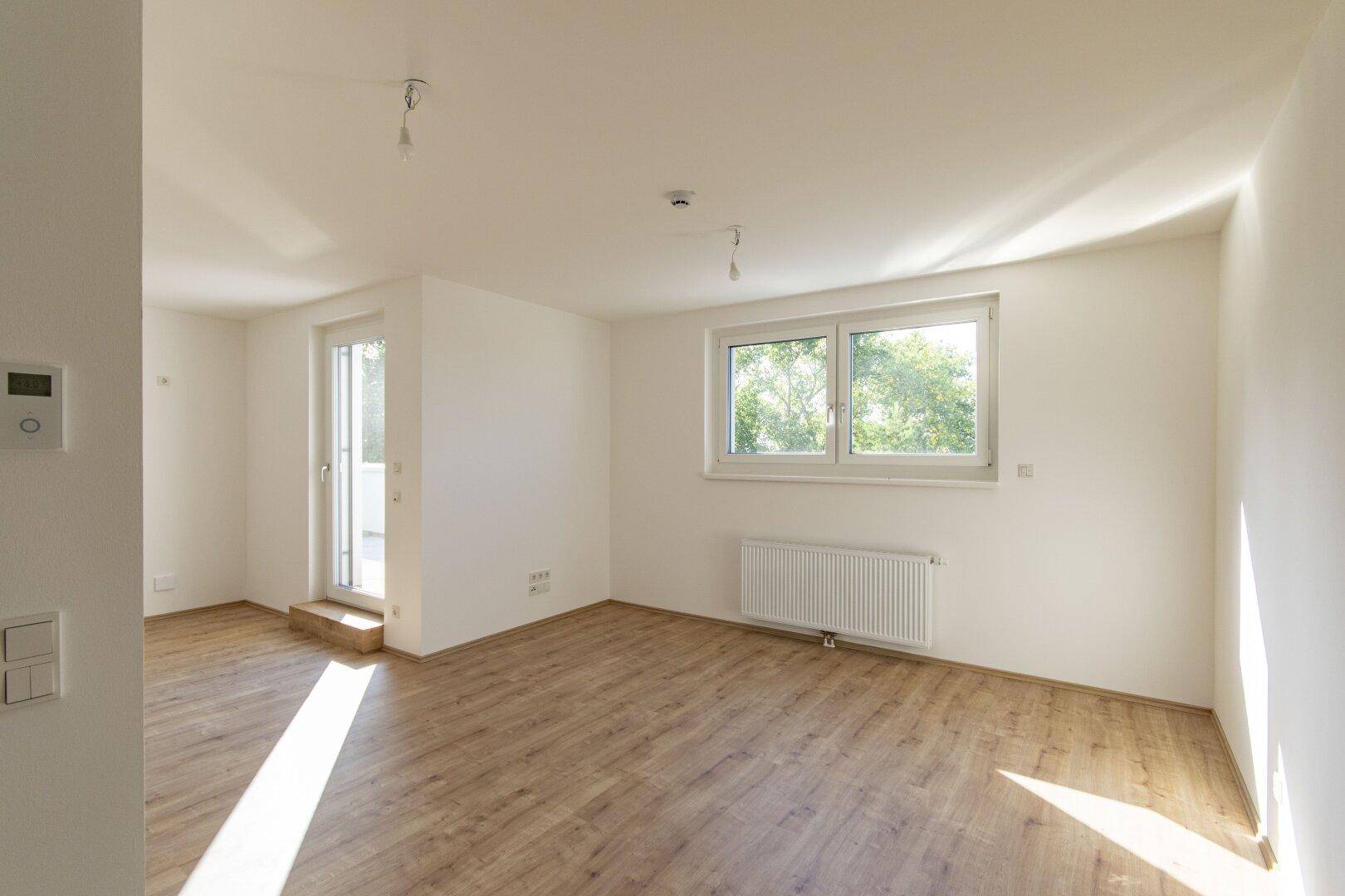 Helles, leeres Wohnzimmer mit Laminatboden, großem Fenster und Zugang zum Balkon.