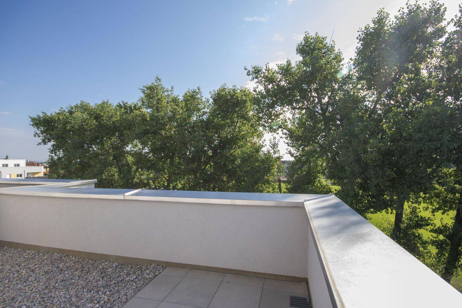 Sonniger Balkon mit Kiesbereich und Fliesenboden, umgeben von hohen Bäumen.