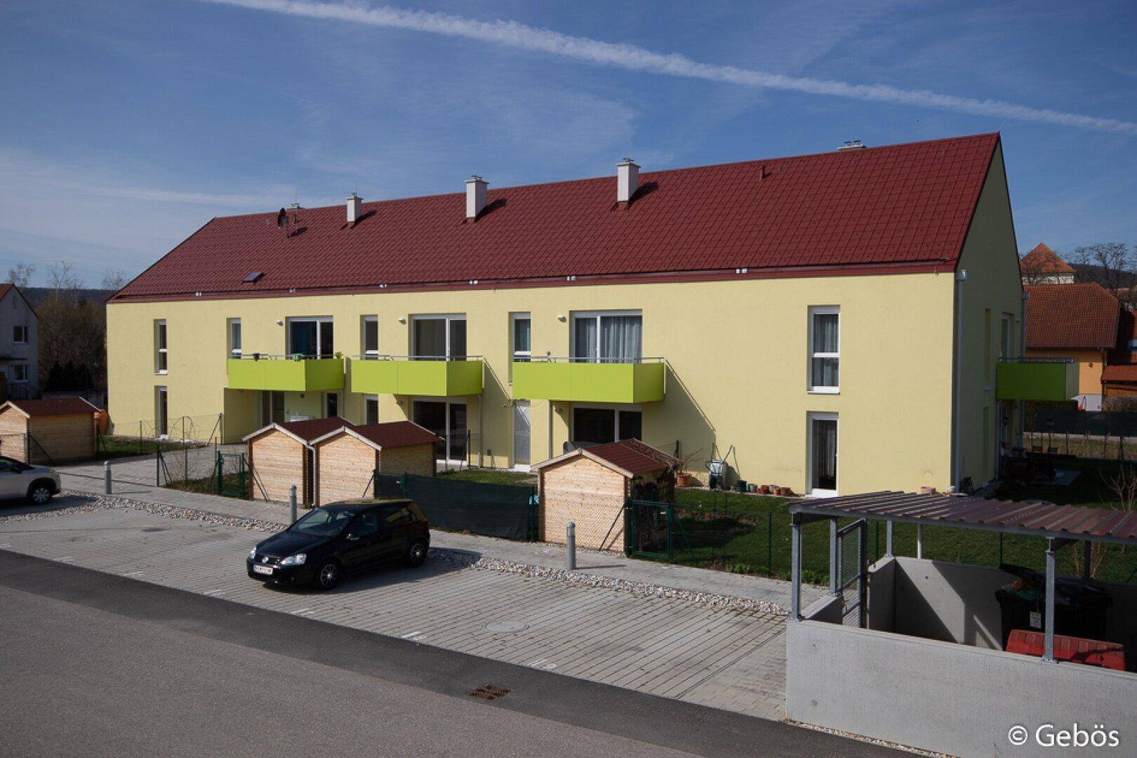 Modernes Mehrfamilienhaus mit gelber Fassade, roten Dächern und grünen Balkonen, inklusive Parkplätzen.
