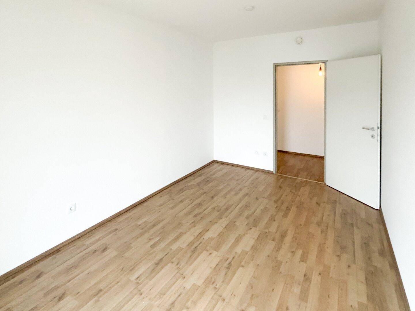 Leeres Zimmer mit hellem Laminatboden und weiß gestrichenen Wänden, bietet vielseitige Nutzungsmöglichkeiten.