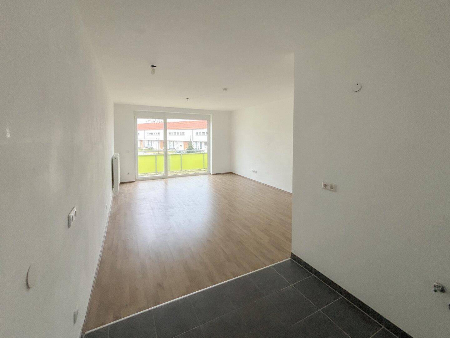 Heller, leerer Wohnbereich mit Laminatboden und Zugang zu einem kleinen Balkon oder Garten.