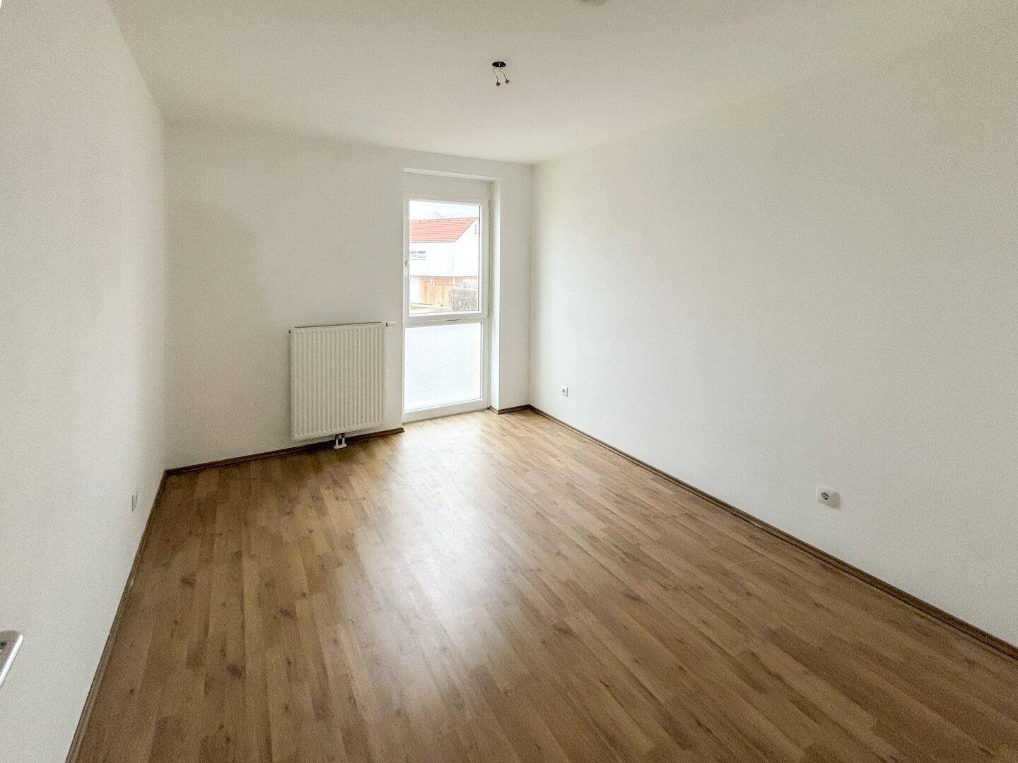 Leeres Schlafzimmer mit Laminatboden, weiß gestrichenen Wänden und einem Fenster mit Heizkörper.