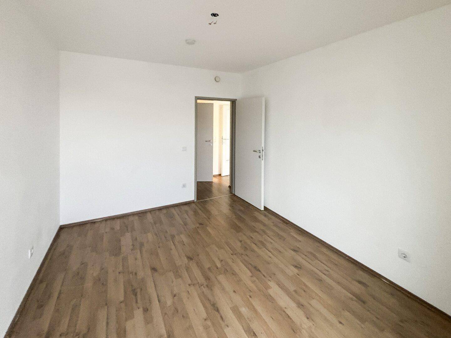 Helles Zimmer mit Laminatboden und weiß gestrichenen Wänden, eine offene Tür führt zum Flur.