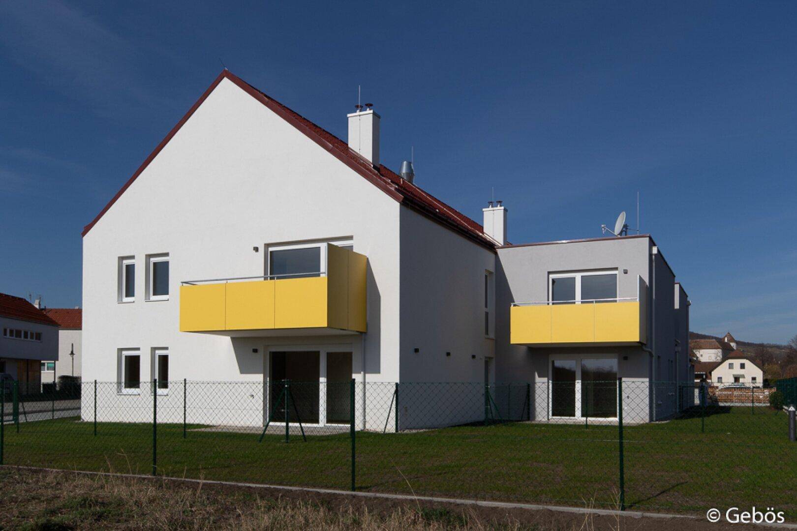 Seitenansicht eines Neubaus mit weißer und grauer Fassade sowie gelben Balkonen.